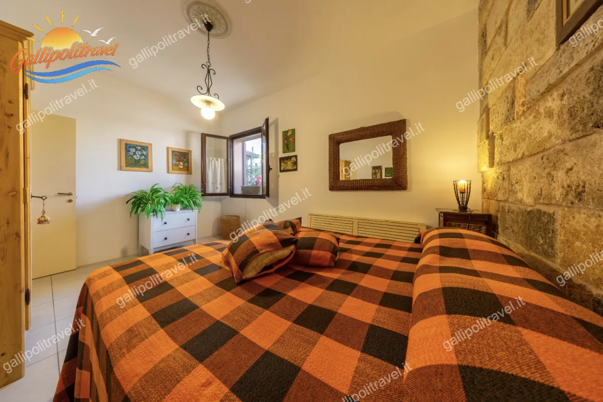 Dettaglio della luminosa camera matrimoniale del Trilocale Green, che mostra la finestra aperta, il letto a scacchi e un comò bianco decorato con fresche piante verd